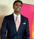 A smiling man in a navy blue suit, white dress shirt, and a pink paisley patterned tie and pocket square, standing in front of a textured beige wall and a bright pink and yellow background.
